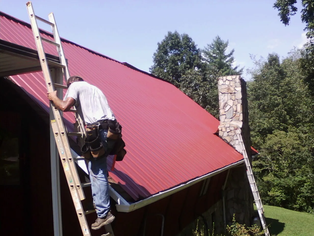 Roofer on a ladder beside a red metal roof and stone chimney — Soffit & Fascia Repair by Roofing Brothers in Waco